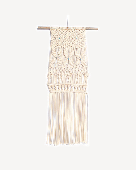 Tenture murale en macramé écru avec perles turquoises et branche de bois naturel.